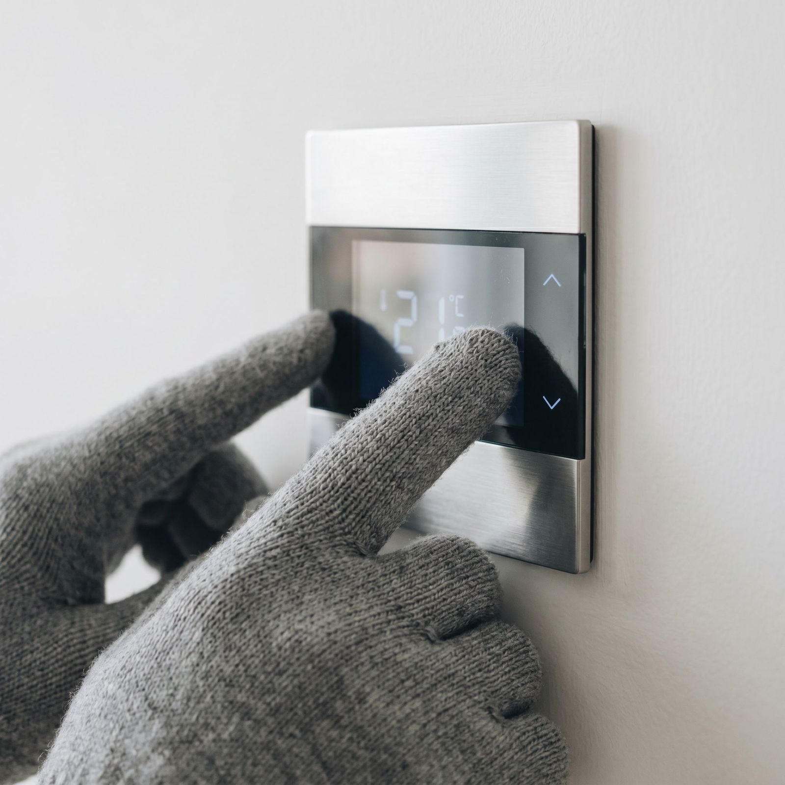 A gloved hand adjusting a wall-mounted digital thermostat.